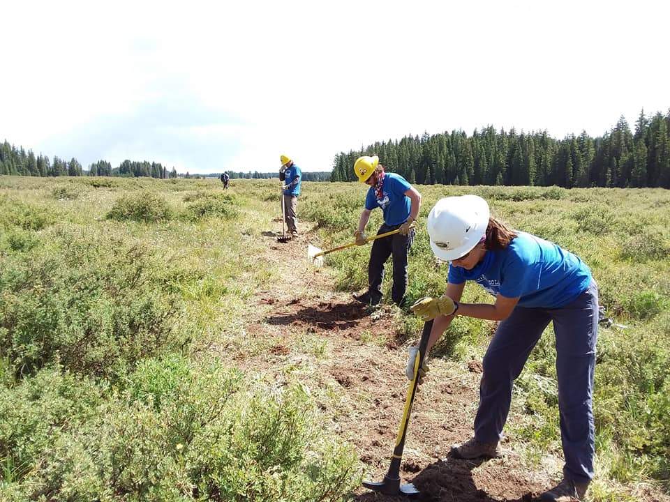 Three people in hard hats doing trail work in an open field with a treeline behind them